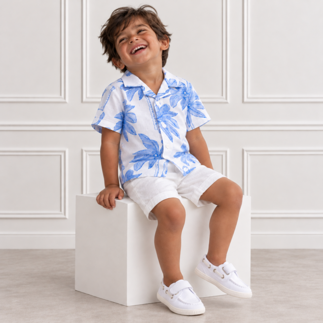 Indian boy laughing joyfully on a white cube, wearing a blue palm print shirt, white shorts and white shoes, in a bright studio.